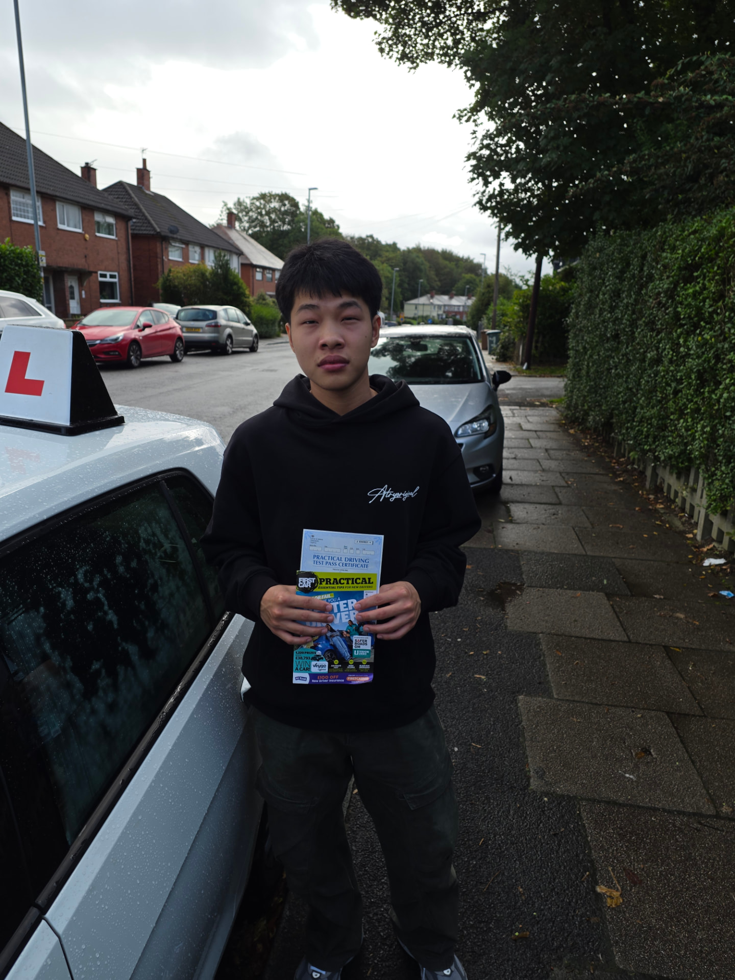 FSK Driving Instructors learner Yixun from Middleton holding his practical driving test pass certificate next to the learner car after passing first time at Chadderton test centre