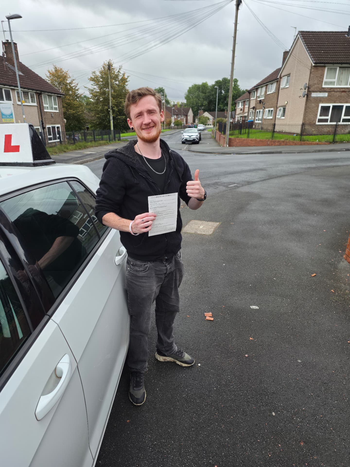 FSK Driving Instructors learner Damien holding his practical driving test pass certificate and giving a thumbs-up next to the FSK learner car after passing his driving test in Rochdale